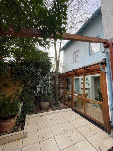 Hinterhof mit Pflanzen, einem Holzrahmen und einem blauen Haus im Hintergrund.