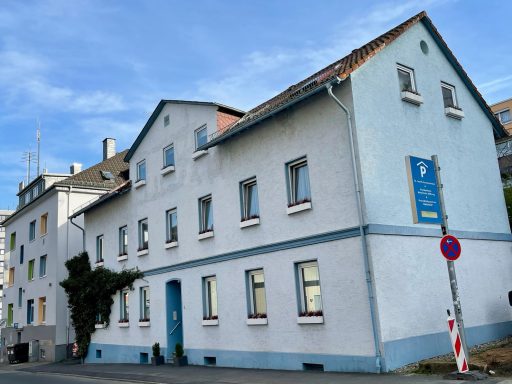 Weißes Gebäude mit blauen Akzenten an einer Straßenecke, darunter ein Schild.