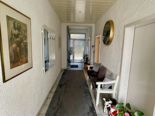 Ein heller Flur mit Bildern, einer Bank und Pflanzen am Boden.