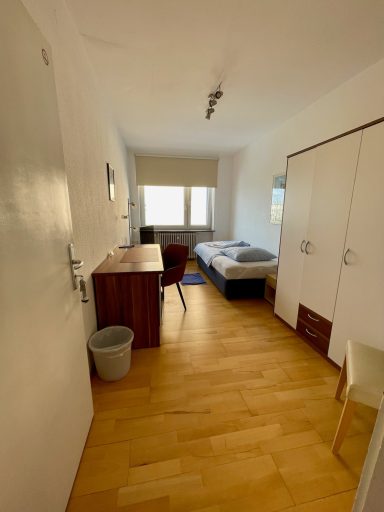 Heller, einfacher Raum mit Bett, Tisch, Stuhl und Schrank. Holzfußboden.