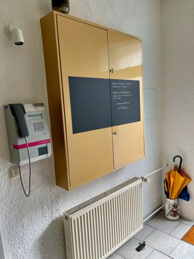 Brauner Schrank an der Wand, neben einer Gegensprechanlage und einem Heizkörper.