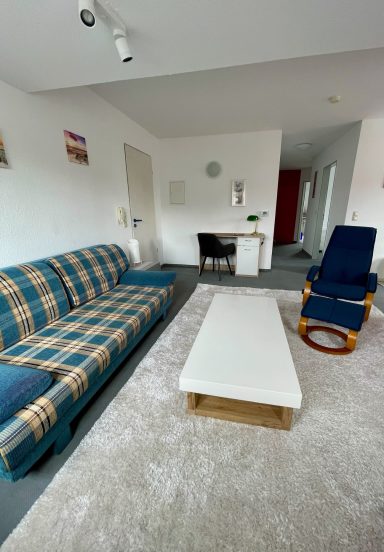 Wohnzimmer mit Sofa, Sesseln, Tisch und Teppich in hellen Farbtonen.