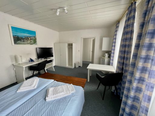Helles Zimmer mit Doppelbett, Schreibtisch, TV, Kühlschrank und Stühlen.